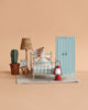 The Maileg Little Sister Bedroom Set features a small mouse on a vintage bed in a miniature room with a blue door, cactus plant, lamp, alarm clock, rug, and red lantern—all arranged against a beige background.