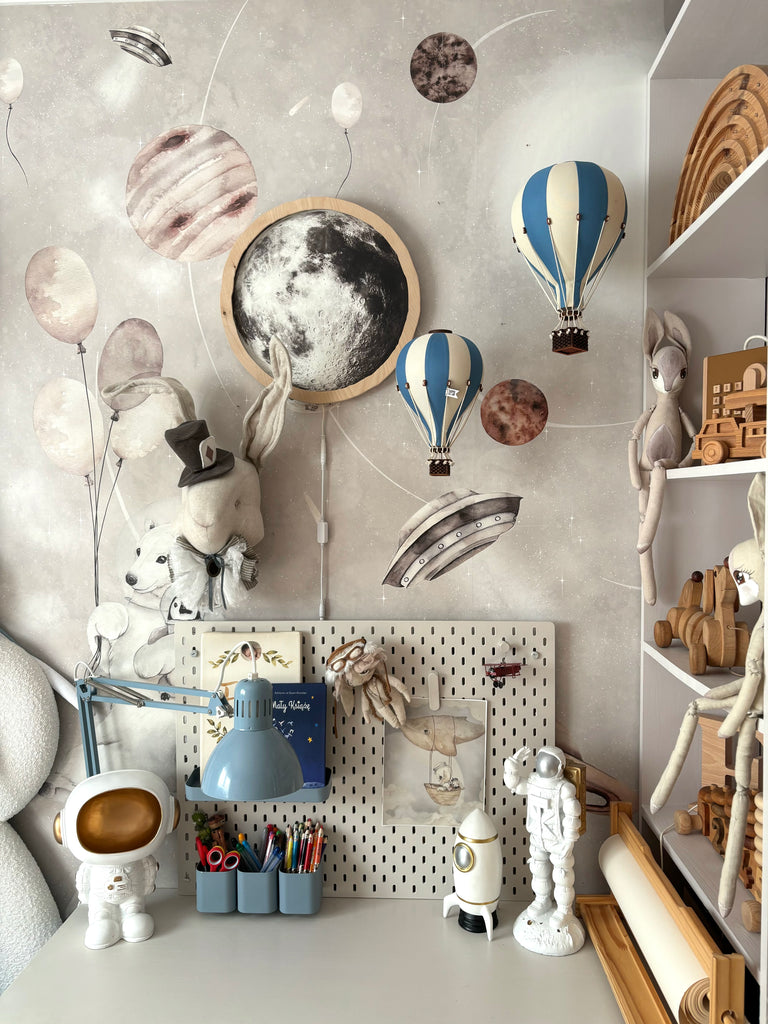 Children’s desk with astronaut figures, blue lamp, art supplies, and wall art featuring a moon, planets, rabbits wearing hats, and blue-striped hot air balloons—complemented by the charming "Hot Air Balloon - Marine Blue" ornament—creating whimsical space-themed handmade children's room decor.