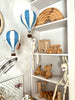 White shelves display wooden toys—such as a truck, phone, rainbow arches, pull-along dog, rabbits, and the letter "F"—while soft, neutral colors create a cozy and whimsical children's space. The room is accented with charming handmade decor and the Hot Air Balloon - Marine Blue, adding an organized and delightful touch to the décor.