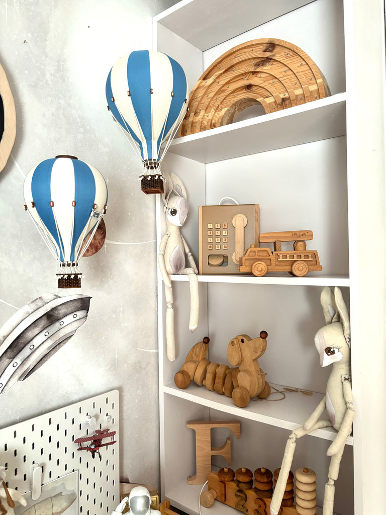 White shelves display wooden toys—such as a truck, phone, rainbow arches, pull-along dog, rabbits, and the letter "F"—while soft, neutral colors create a cozy and whimsical children's space. The room is accented with charming handmade decor and the Hot Air Balloon - Marine Blue, adding an organized and delightful touch to the décor.
