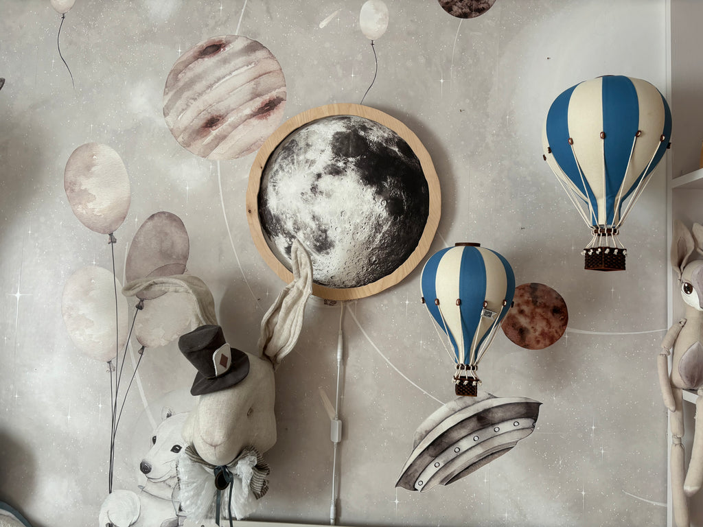 A whimsical wall decor featuring the "Hot Air Balloon - Marine Blue" plush, accompanied by a charming rabbit in a hat, blue and white wooden balloon ornaments, planets, and a moon illustration set against a starry background—perfect as handmade children's room decor.