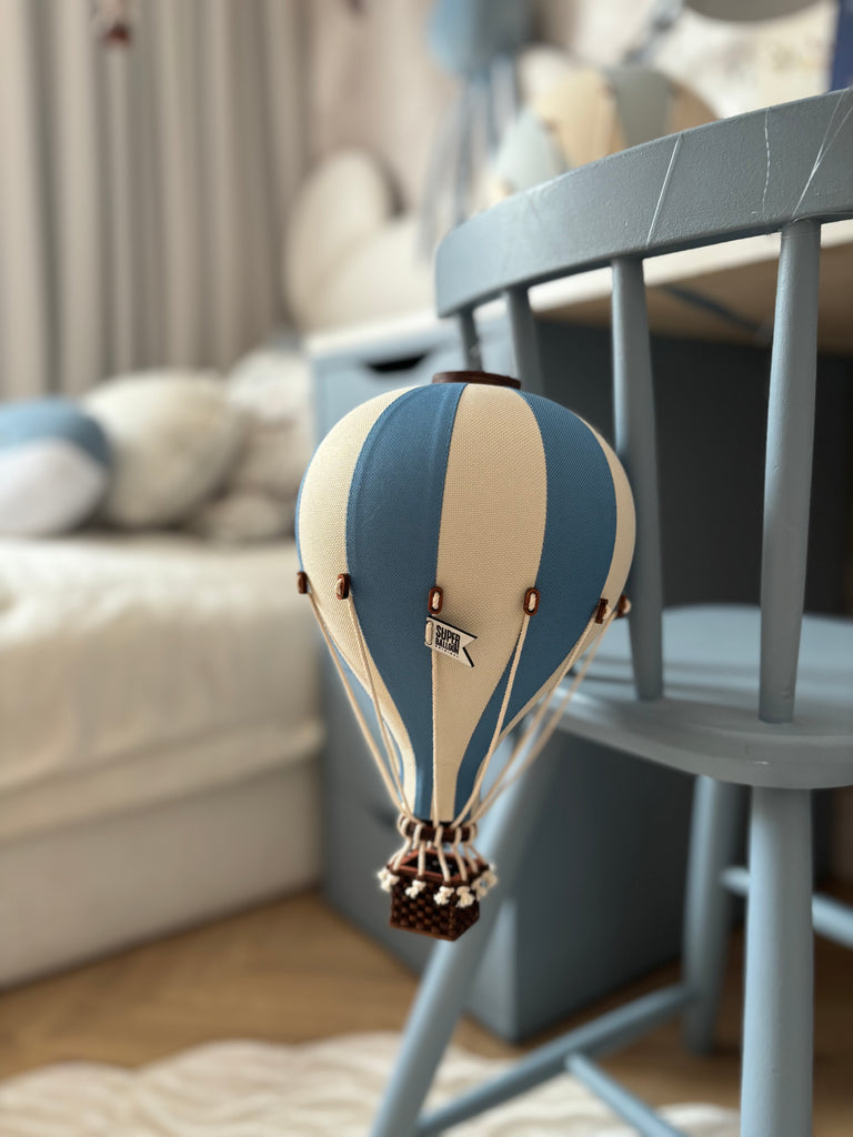 A small decorative Hot Air Balloon - Marine Blue wooden ornament hangs from a chair in a cozy, softly lit room with a bed and other furniture in the background.