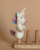 The Unicorn Finger Puppet is an adorable, small felt toy featuring a white body, a pink nose, and a mane and tail in shades of blue, purple, and pink. This needle-felted unicorn has a white horn and is perched on a wooden stand with three pegs. The background is plain beige.