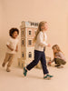 Three children play around the Fully Furnished Dollhouse With Lights, House of New York - The Six-Storey Giant. Two kids run and smile, while one kneels beside the dollhouse, opening a door. The background is beige and minimalistic.
