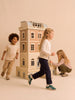 Three children play around the Fully Furnished Dollhouse With Lights, House of New York - The Six-Storey Giant. One child runs in the foreground, another laughs while walking, and a third kneels beside the intricately detailed, fully furnished dollhouse, engaging with its features. The background is a light beige color.