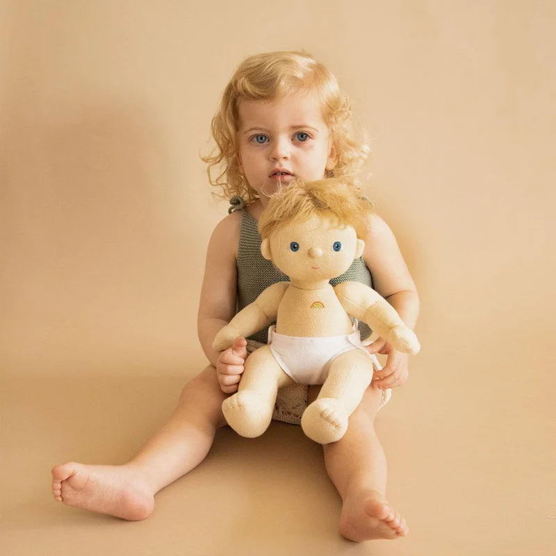 A toddler with curly hair sits on a beige backdrop, holding a Dinkum Doll - Poppet (Extended Pack), looking thoughtfully at the camera. Both the child and doll are in light, neutral-toned clothes with removable pieces.