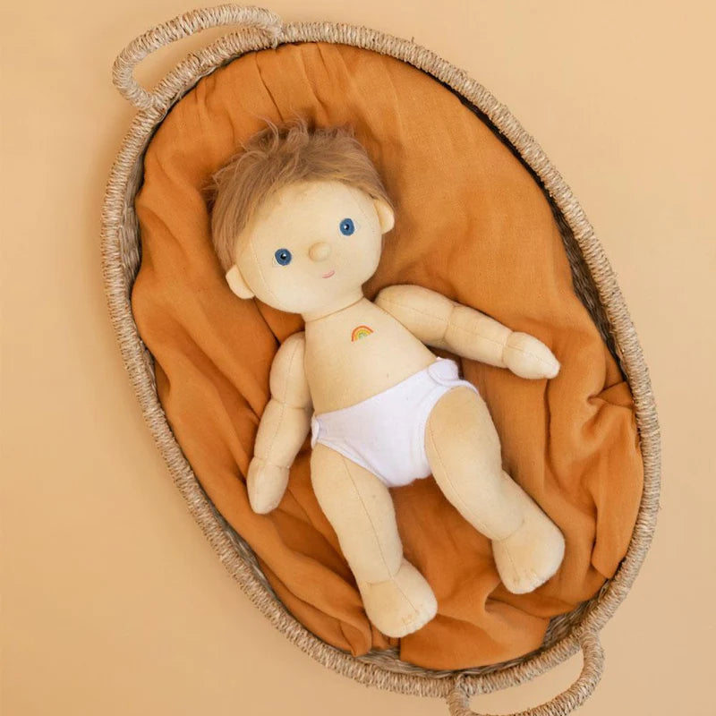 A plush Dinkum Doll - Poppet (Extended Pack) with blue eyes and brown hair, lying on a mustard yellow cloth inside a half-moon shaped basket, wearing a removable unisex outfit.