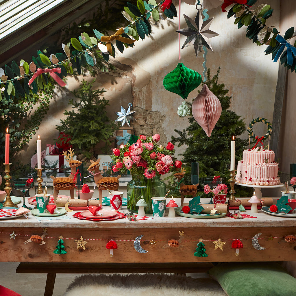 The festive holiday table, adorned with red and pink flowers and twinkling candles, showcases a layered cake. MeriMeri Large Hanging Decorations and luxurious ornaments contribute to the seasonal cheer. Christmas trees and garlands further enhance the warm, inviting backdrop.