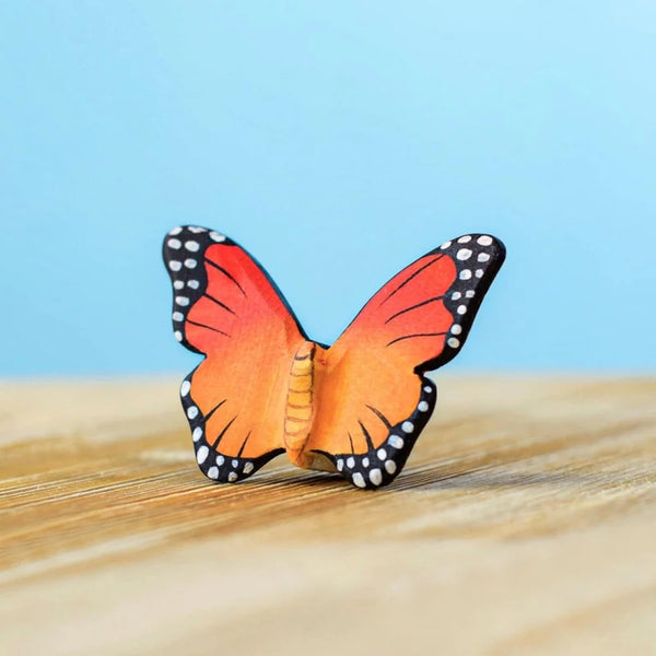 The Bumbu Handmade Wooden Orange Butterfly, a small handcrafted toy with vibrant orange and black wings adorned with white spots, is shown on a wooden surface against a light blue background.