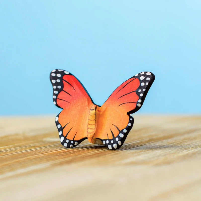 The Bumbu Handmade Wooden Orange Butterfly, a small handcrafted toy with vibrant orange and black wings adorned with white spots, is shown on a wooden surface against a light blue background.