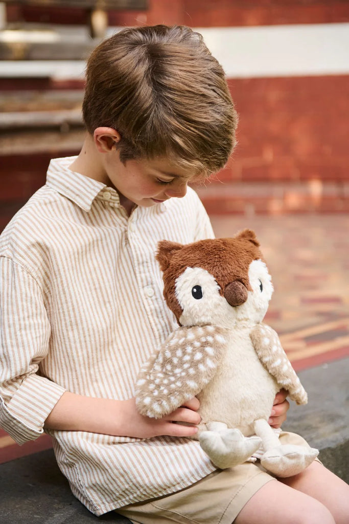 A young boy, brown-haired and dressed in a striped shirt and shorts, sits on a bench holding Oscar The Owl Stuffed Animal, blending into the bohemian outdoor scene with a brick wall as the backdrop.