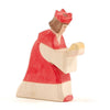 A handcrafted wooden figurine depicting Ostheimer Red King With Crown, wearing a red and white outfit, focused on reading a book. The figure is simplistic, with minimalistic details and a visible wood grain.