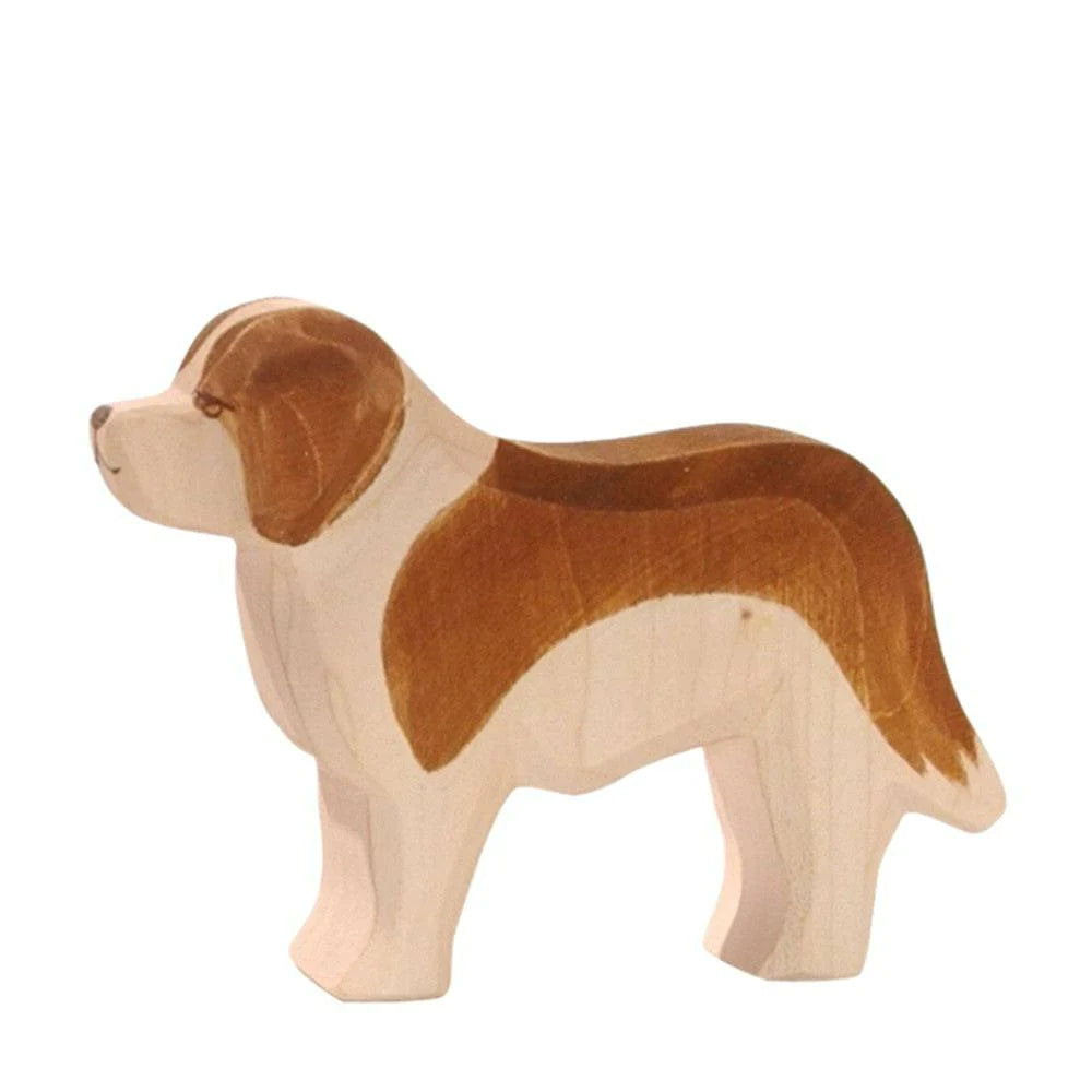 A handcrafted wooden figurine of a dog with brown and white markings. The dog is in a standing position, carved in a simple, stylized manner. The wood grain and texture are visible, highlighting its artistic craftsmanship, reminiscent of the Ostheimer Saint Bernhard Dog perfect for imaginative play.
