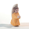 A hand-painted wooden figurine of a girl, depicted with brown hair and a yellow dress, set against a plain white background. This Ostheimer Little Angel - Orange is stylized and simplistic, capturing a