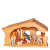 A wooden nativity scene featuring figurines of the holy family, three animals, and an angel under a Ostheimer Stable Barn structure, all handcrafted in simple, colorful styles against a white background.