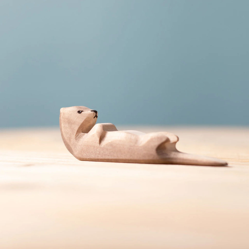 The Bumbu Handmade Wooden Otter Sleeping figure rests on its back atop a light wooden surface with a soft blue background, highlighting its eco-friendly craftsmanship.