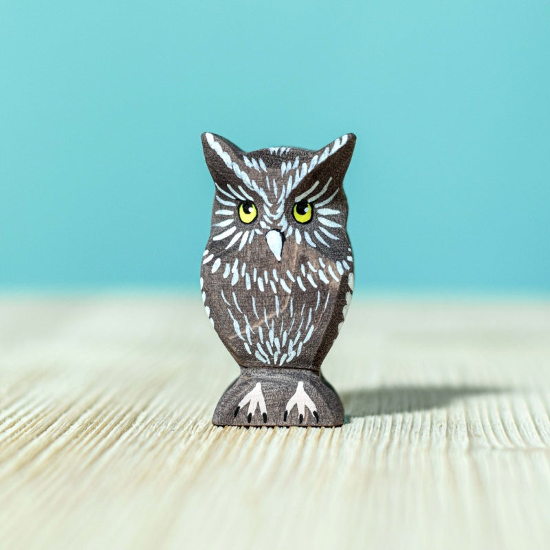 The Bumbu Handmade Wooden Ancient Oak Tree with Owl Set features a small painted owl with yellow eyes, crafted from sustainable maple wood, standing on a light wooden surface against a solid light blue background.