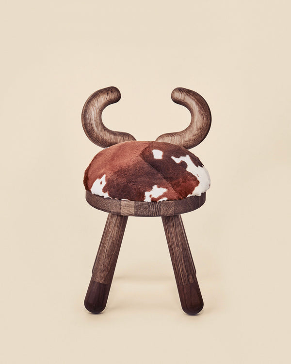 A unique children's Faux Cow Chair with dark wooden legs and a cushioned seat upholstered with faux fur, designed to resemble horns at the top. The background is a soft beige.