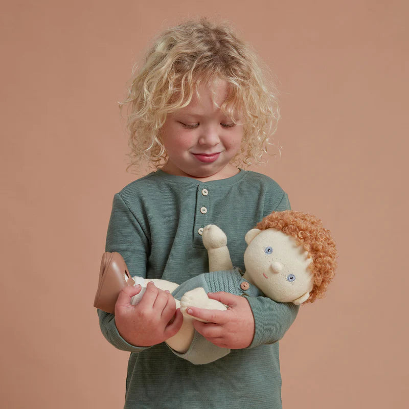 A young child with curly blond hair, wearing a green shirt, gently holds a broken ceramic bird and an Olli Ella | Dinkum Doll - Pea, standing against a soft peach background.