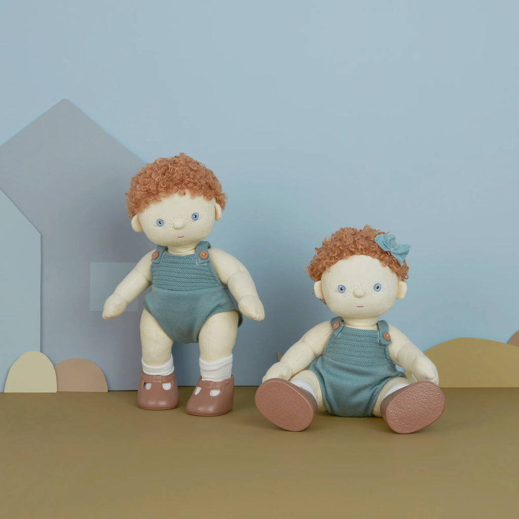 Two Olli Ella Dinkum Dolls with curly red hair and green overalls sitting against a pastel blue backdrop, with cut-out paper shapes decorating the scene.