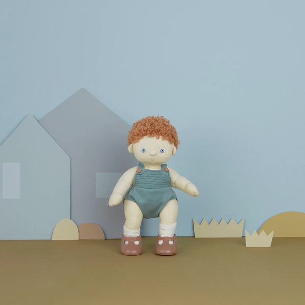 A Olli Ella | Dinkum Doll - Pea with curly red hair and blue overalls stands in an imaginative playtime set depicting a town, with paper cutouts of blue houses and brown hills against a beige background.