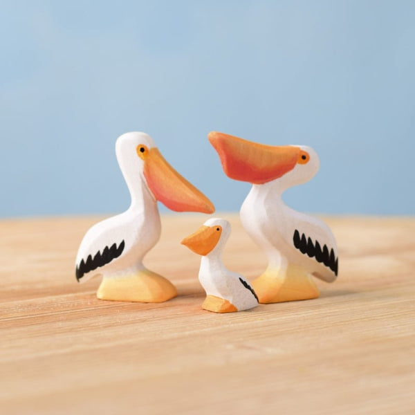 The Bumbu Handmade Wooden Pelicans Family Set features three white wooden pelican toys with black and orange details, arranged on a light wooden surface against a blue background—ideal for Montessori play and sustainable toy collections.