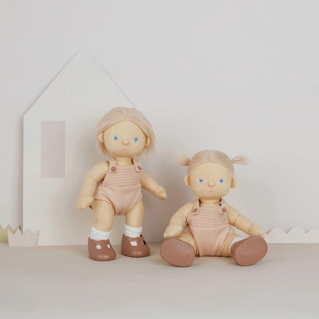 Two plush, posable Olli Ella Dinkum Dolls in Petal with blonde hair, dressed in pink overalls, are set against a neutral background with minimalist decor. One doll is standing while the other is seated.