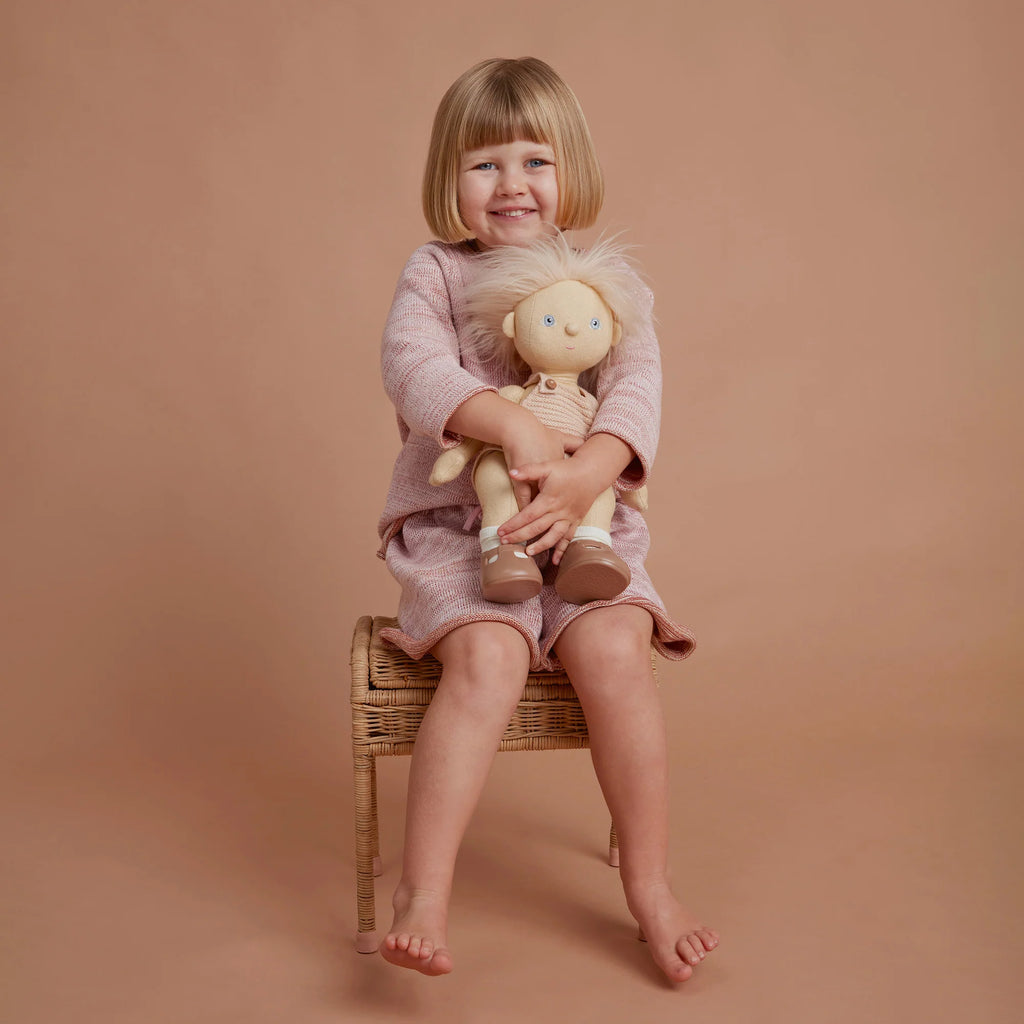 A young girl with blonde hair, dressed in a pink dress and cardigan, sits on a wicker stool hugging an Olli Ella | Dinkum Doll - Petal, against a soft peach background.