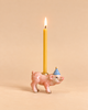 A delightful pink ceramic Pig Cake Topper wears a blue and white striped party hat and carries a lit yellow candle on its back, set against a beige background. This charming, hand-painted decoration makes an ideal collectible keepsake for special celebrations.