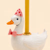 The Pink Goose Cake Topper - Final Sale is a handcrafted white ceramic figurine featuring a goose wearing a pink polka dot party hat and pink bow tie, elegantly holding a yellow candle on its back. This delightful porcelain decoration doubles as a unique cake topper and makes a charming collectible keepsake for special occasions.