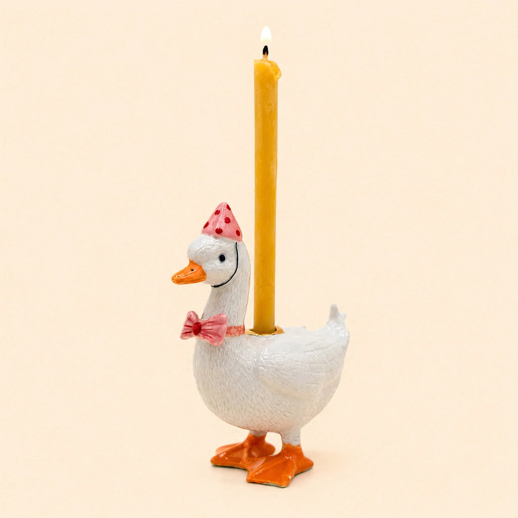 Introducing the Pink Goose Cake Topper – Final Sale: a handcrafted birthday decoration shaped like a white goose wearing a pink party hat and bow tie, elegantly holding a lit yellow candle on its back, set against a light beige background.