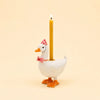 The Pink Goose Cake Topper features a charming white goose adorned with a pink party hat and bow tie, carrying a lit yellow candle on its back. Set against a plain beige background, this handcrafted porcelain decoration makes for a delightful and whimsical cake topper, perfect for birthdays.