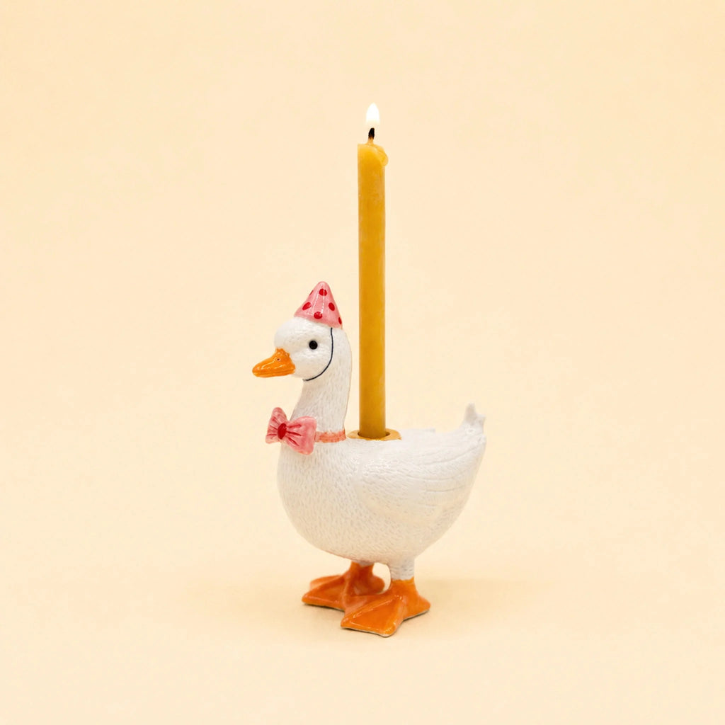 The Pink Goose Cake Topper features a charming white goose adorned with a pink party hat and bow tie, carrying a lit yellow candle on its back. Set against a plain beige background, this handcrafted porcelain decoration makes for a delightful and whimsical cake topper, perfect for birthdays.
