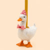 The Pink Goose Cake Topper stands upright with a white body, wearing a pink party hat adorned with red dots and a matching pink bow tie featuring a red heart. A yellow candle rests on its back, all set against a pale background—an adorable handcrafted piece perfect as a collectible birthday keepsake.