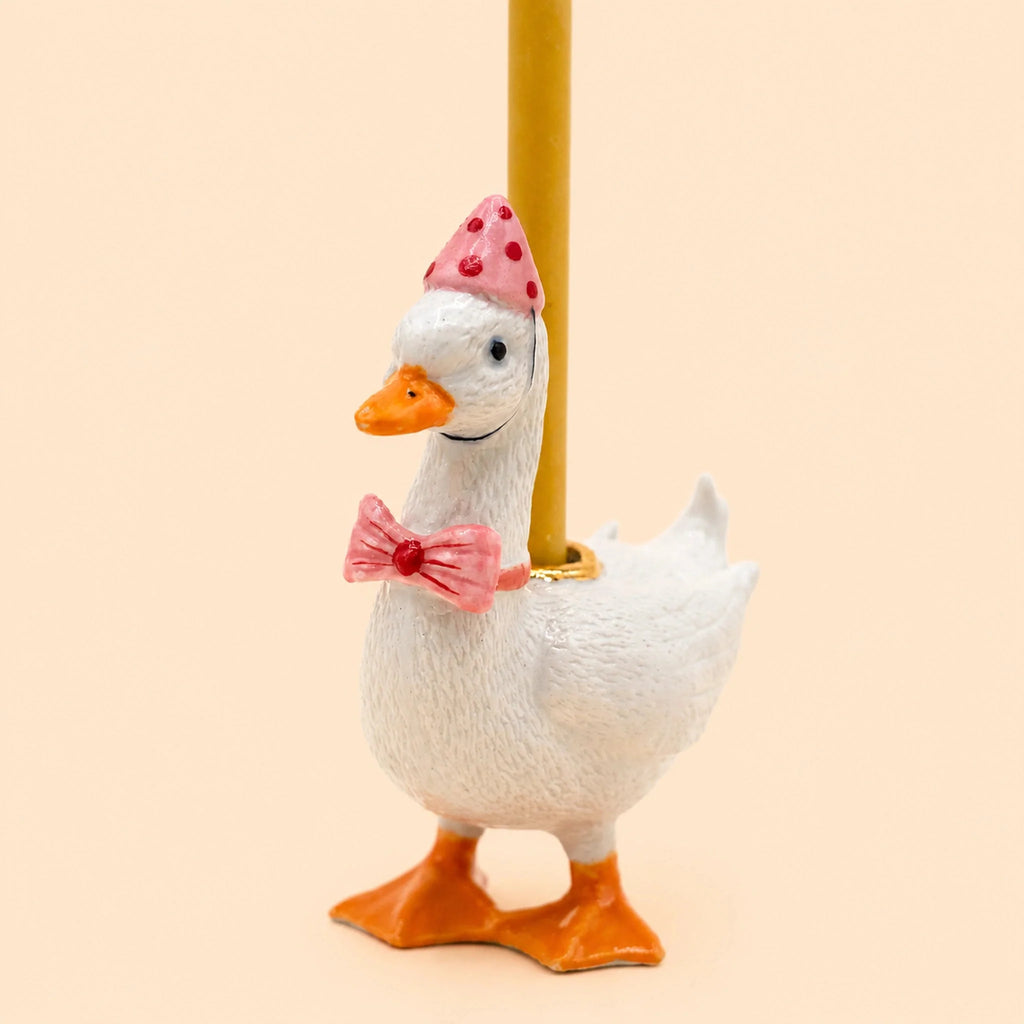 The Pink Goose Cake Topper stands upright with a white body, wearing a pink party hat adorned with red dots and a matching pink bow tie featuring a red heart. A yellow candle rests on its back, all set against a pale background—an adorable handcrafted piece perfect as a collectible birthday keepsake.