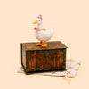 The Pink Goose Cake Topper—now available for final sale—is a charming handcrafted birthday decoration featuring a decorative ceramic goose adorned with a pink bow and polka-dot hat, poised atop a vintage wooden chest on a beige star-patterned cloth, set against a light background; it’s the perfect porcelain accent to crown any special celebration.