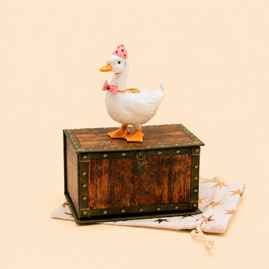 The Pink Goose Cake Topper—now available for final sale—is a charming handcrafted birthday decoration featuring a decorative ceramic goose adorned with a pink bow and polka-dot hat, poised atop a vintage wooden chest on a beige star-patterned cloth, set against a light background; it’s the perfect porcelain accent to crown any special celebration.