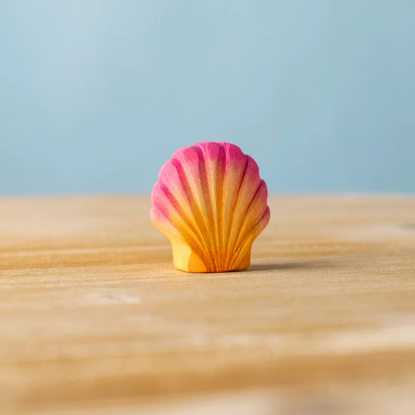 The Bumbu Handmade Wooden Pink Seashell, a small seashell-shaped toy featuring a pink and yellow gradient, sits on a wooden surface against a light blue background.