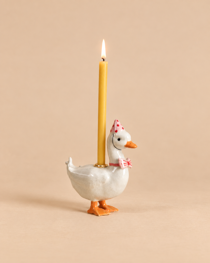The Pink Goose Cake Topper—crafted from white ceramic and hand-painted with care—features a goose wearing a pink polka-dot party hat and bow, holding a lit yellow candle on its back against a simple beige background. This charming collectible is the perfect hand-painted birthday decoration and keepsake.