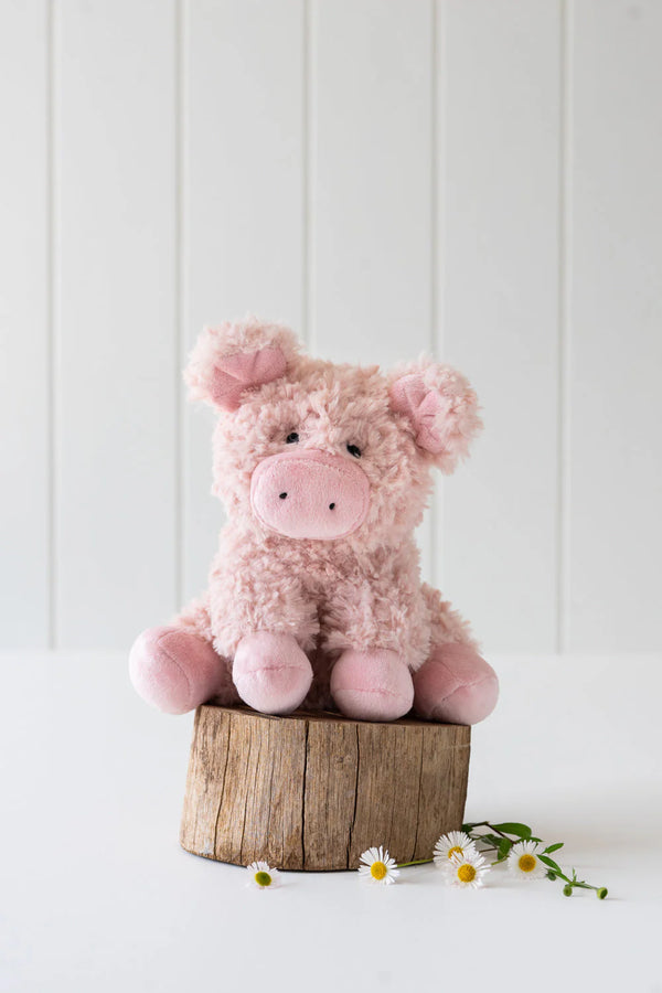 Poppy The Pig Stuffed Animal is perched on a small wooden stump against a white background, echoing *Animal Farm*, with a few small white flowers scattered nearby.