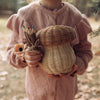 A child in a pink sweater holds an Olli Ella Mushroom Basket and a bundle of twig pencils in a forest setting.