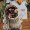 A rusting Olli Ella Mushroom Basket holding cinnamon sticks, adorned with a twine bow and a small floral decoration, placed on a wooden table surrounded by loose flowers and twig pencils.
