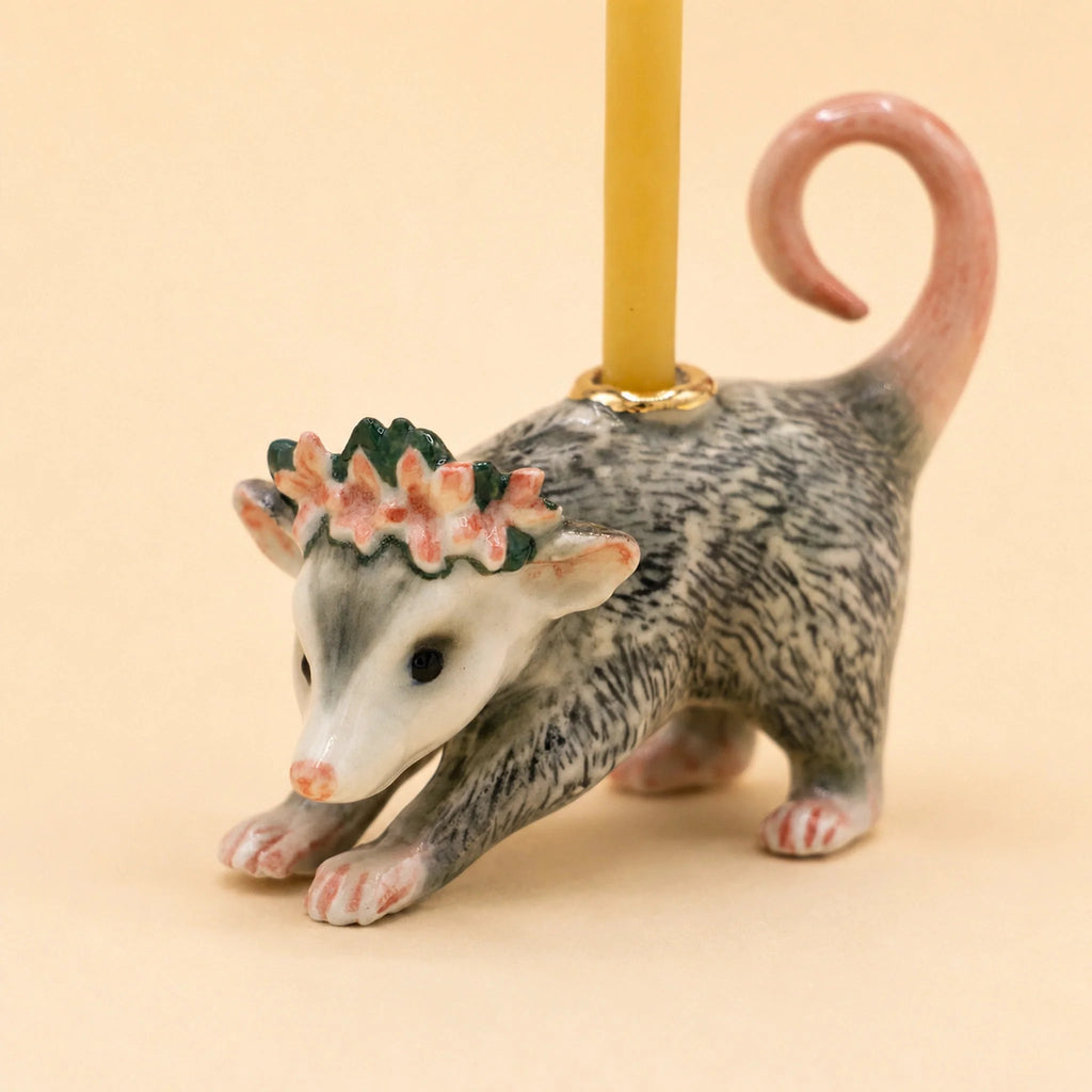 The Possum Cake Topper - Final Sale features a hand-painted birthday candle nestled on the back of a ceramic possum figurine adorned with a flower crown and curled tail, creating a delightful porcelain cake topper and collectible keepsake set against a beige background.