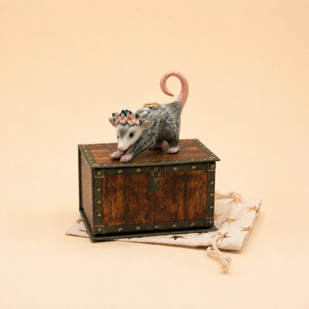 The Possum Cake Topper – Final Sale features a petite possum figurine adorned with a flower crown and balancing a coin on its back, standing atop a wooden chest that rests on a star-patterned cloth bag—an enchanting porcelain keepsake and delightful collectible for any celebration.