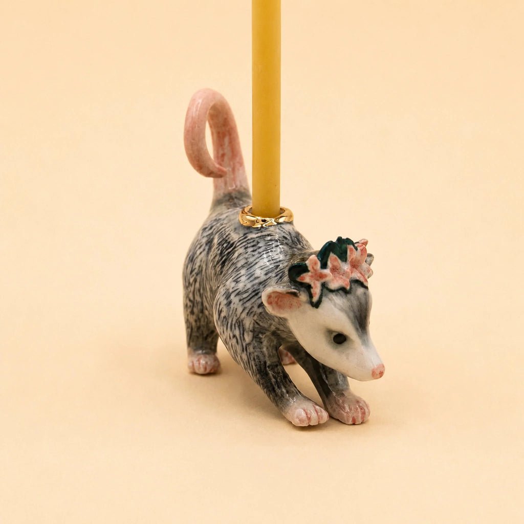 The Possum Cake Topper - Final Sale is a charming, hand-painted ceramic figurine featuring a possum with a pink tail and floral crown, holding a yellow candle on its back—perfect as a birthday candle holder or collectible keepsake, shown against a beige background.