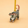 A collectible keepsake, the Possum Cake Topper features a ceramic possum figurine adorned with pink flowers on its head, carrying a yellow stick reminiscent of a hand-painted birthday candle on its back, all set against a pale beige background.