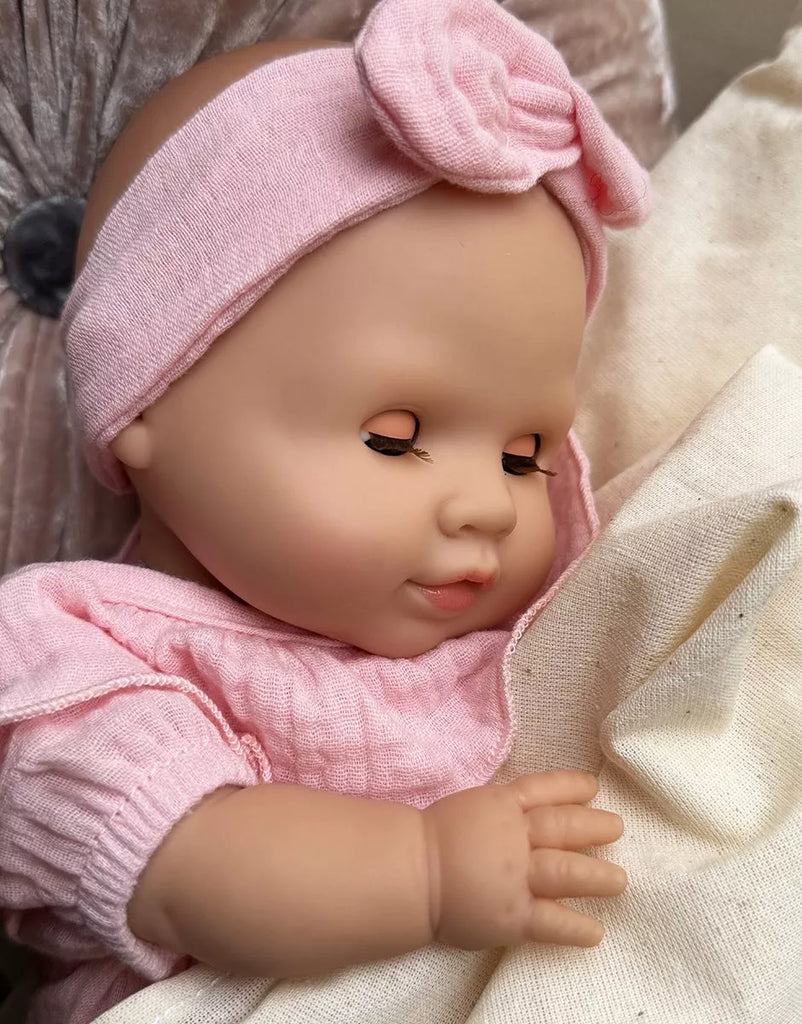 A close-up of the Minikane Cuties Soft Body Doll (14") - Emilie, with closed eyes, wearing a pink outfit and bow headband, gently holding a cream blanket and displaying adorable accessories.