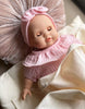 The Minikane Cuties Soft Body Doll (14") - Emilie, with closed eyes and dressed in a pink outfit and headband, lies on cream sheets with a pleated pillow—ideal for styling with your favorite doll accessories.