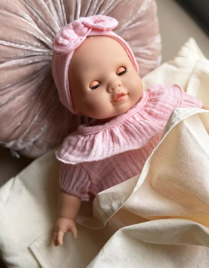 The Minikane Cuties Soft Body Doll (14") - Emilie, with closed eyes and dressed in a pink outfit and headband, lies on cream sheets with a pleated pillow—ideal for styling with your favorite doll accessories.
