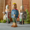 The Bumbu Handmade Wooden Prince, in a blue shirt, green pants, red cape, and yellow crown, stands before two armored guards with a medieval castle toy backdrop—an ideal set to inspire imaginative play.
